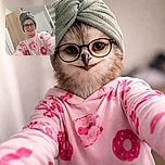 A smiling child wearing glasses and a towel wrapped around their head takes a selfie in a casual room, sporting a pink shirt with red donut and dessert prints, with a slightly tilted wardrobe and shelves in the background.