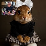 A charming baby wearing a large white bow headband and a stylish black and white dress sits on a colorful blanket on a couch, smiling with wide eyes and tiny hands clasped together, with a baby bottle nearby and a cozy home background.