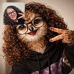 A cheerful young person with curly hair and glasses flashes a peace sign while smiling warmly at the camera against a plain wall background.
