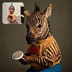 A toddler with a tiny ponytail on top of their head stands on carpeted floor holding a brush, wearing a Mickey Mouse t-shirt and blue shorts, looking curiously at the camera in an indoor setting with a white door and storage unit in the background.