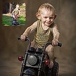 A cheerful toddler is posing on a tiny black ride-on motorcycle, looking like the world’s smallest biker gang leader while cruising through the backyard. Bright sunlight, green grass, rocks, garden furniture, and a yellow toy wagon complete the playful outdoor scene.