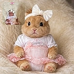 A sweet baby girl dressed in a delicate pink and white outfit with a big bow headband, lying on a fluffy white blanket with a '3 months' sign above her, looking curiously at the camera.