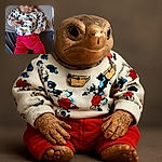 A cute baby dressed in a Mickey Mouse themed sweater and bright red pants lounges comfortably on a cushion with a woven white backdrop, looking curiously at the camera with a slight pout and tiny hands in motion.