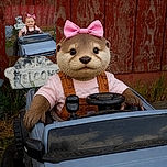 A smiling baby with a spectacular pink bow is cruising a toy pickup truck like the tiniest off-road champion, complete with steering wheel, gear shifter, and a watchful garden bear co-pilot.