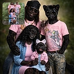 A cheerful family portrait where everyone coordinated on pink Tom & Jerry shirts like it was a themed superhero squad — two grown-ups standing protectively, a smiling mom seated, and an adorable baby with a bow stealing the spotlight. Studio green-bokeh backdrop and wood floor complete the very Instagram-ready vibe.