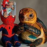 A curious baby is sitting in a colorful baby seat, holding a blue object close to their mouth with wide eyes, next to a knitted Spiderman doll that looks ready to save the day.