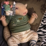 A tiny baby lounges like a pint-sized celebrity on a patterned blanket, wearing a blue cap, dinosaur shirt, and cartoon pajamas while flashing an impressively serious expression and a little raised-fist pose.