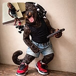 A joyful young child with beaded hair strikes a playful pose against a plain wall, sticking out their tongue and holding a phone, rocking bright red sneakers and casual shorts with infectious energy.