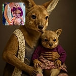 A smiling woman kneels on the grass holding a wide-eyed baby dressed in a purple top and floral skirt, with the baby perched on her knee. The scene is outdoors with other people partially visible in the background, and the woman wears a white knitted vest and patterned dress.