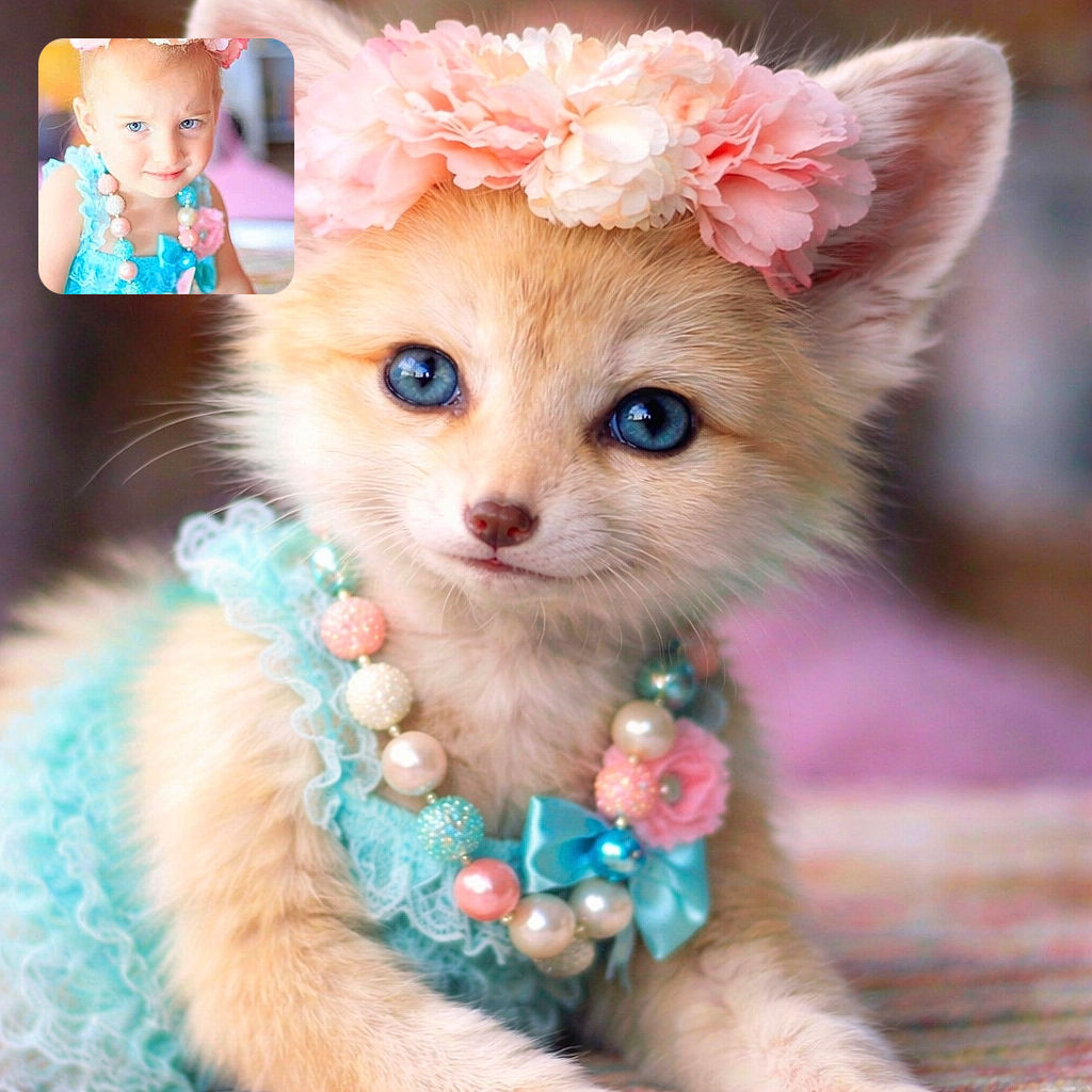 A charming young child wearing a blue dress and a pink floral crown, adorned with pastel bead necklaces, looks playfully at the camera with a slight smile. The background is softly blurred, emphasizing the child's bright blue eyes and delicate features. The photo has a watermark text overlay from the photographer.