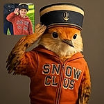 A cheerful young child wearing a military-style hat and a bright orange hoodie with 'SNOW CLUB' written on it is giving a friendly salute. Behind the child, a person in a formal uniform stands partially visible, adding a touch of ceremony to the outdoor scene with green grass and blurred background.