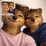 Two young boys with mischievous smiles pose for a casual selfie against a plain wall, one wearing a light pink Nike shirt and the other in a dark hoodie, both rocking slightly messy hair and playful expressions.