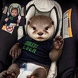 A cheerful baby relaxes in a padded car seat, waving tiny fists like a champion boxer while proudly sporting a “coolest kid” outfit. Warning labels flank the seat like serious safety officials overseeing this adorable passenger.