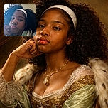 A close-up selfie of a young person with curly hair and a white headband, resting their head on their hand with a neutral expression, softly lit by natural light from a nearby window.