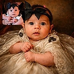 A wide-eyed baby with a big black bow on her head is being gently held by an adult, whose face peeks lovingly from behind. The baby looks curious and slightly surprised, with chubby arms and tiny hands gripping the adult's fingers tightly. The warm lighting highlights the soft skin and tender moment between the two.