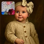A charming baby wearing a cozy cream knit sweater and a large bow headband lies comfortably on a textured surface next to a vibrant bouquet of red roses and greenery, looking off to the side with curious, sparkling eyes.