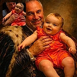 A joyful baby in a bright red outfit is being held up by a smiling man against a black background, capturing a warm and happy moment filled with love and playful energy.