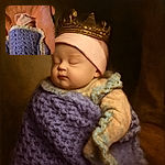 A peaceful sleeping baby bundled up in a cozy pink hat and wrapped snugly in a handmade purple crocheted blanket, lying on a soft peachy textured surface.