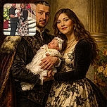 A proud couple stands indoors by a floral arrangement and steps, the man holding a peacefully sleeping baby dressed in white and yellow, while the woman smiles warmly in a floral dress and black cardigan, creating a cozy family moment in a softly lit setting.