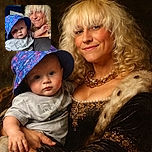 A cheerful adult with curly blonde hair and a black hat smiles warmly while holding a curious baby wearing a colorful blue bucket hat and a gray shirt, both posing indoors with soft lighting highlighting their faces.