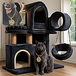 A regal gray cat wearing a large gold chain pendant is being gently held by a person dressed in black. The cat looks calm and slightly unimpressed, set against a cozy indoor background with a sun and moon tapestry and wooden furniture.