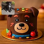 A fluffy brindle puppy with white paws and a green collar sits adorably on a wooden floor next to a bed with a green and white patterned blanket, looking curiously at the camera with a calm expression.
