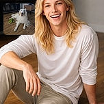 A fluffy white dog with tan patches lounges happily on a wooden floor next to a green plush toy frog, surrounded by a cozy room with a cardboard box and dark shelving unit in the background.