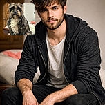A scruffy, curious dog with a charming beard and expressive eyes sits attentively on a wooden surface in a cozy room, framed by a classic wooden cabinet and a comfy bed with colorful blankets.