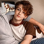 A tiny, adorable puppy with a brown and white coat is comfortably nestled on a person's lap, looking up with soulful eyes and a slightly tilted head, wearing a brown collar. The cozy setting includes denim jeans and a gray zip-up jacket, suggesting a warm, affectionate moment during a car ride.