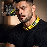 A large black dog with a shiny coat and a bright yellow collar sits on a couch in a cozy room. The dog looks thoughtful or slightly sad, with one ear flopped back. In the background, a cushioned chair and a window with closed blinds and curtains add to the homely atmosphere.