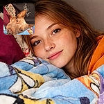 A sleepy brown dog burrowed under colorful fleece blankets stares up like it was rudely woken for portrait duty — one ear proudly erect, nose nearly touching the camera, with a patterned comforter and a fan peeking in the background creating a cozy, chaotic bedroom vibe.