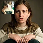 A regal tabby and white cat lounges with its paws draped over a textured green surface, staring intently with calm green eyes. The soft light highlights its fluffy fur and the patterned fabric in the background adds a cozy touch.