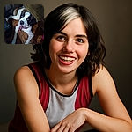 A happy dog with a black and white face and a pink harness is lying on a wooden floor next to a brown couch, looking up with an inviting smile and bright eyes, creating a cozy indoor vibe.
