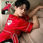 A stylish black and white cat lounging on a beige blanket, dressed in a bright red outfit with a heart that says 'XOXO', giving off major Valentine vibes and a look of mild curiosity.