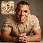 A happy dog lounges on a patch of dry grass with its tongue out, enjoying a sunny day near a blurred natural background of trees and water.