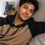 A goofy big-eared dog flopped in the corner, giving the camera a sleepy side-eye with its tongue peeking out — looks part model, part houseplant. The warm brown and black coat fills the frame while a plain white wall and tile floor keep the vibe minimal and very cozy.