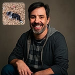 A happy black and white dog sits among a thick carpet of brown leaves, while a person is almost completely buried under the leaves with only their hands, shoes, and a colorful hat visible, creating a playful autumn scene.