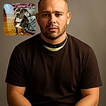 A charming dog with a curious expression sits attentively on a wooden floor next to a cozy pile of blankets, looking like it just heard the word 'treat'. The warm indoor lighting adds a homey vibe to the slightly blurry but heartfelt snapshot.