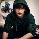 A sleek black cat with striking green eyes is wearing a snug green hoodie, sitting alertly on a soft, cozy blanket. The background is a softly blurred room with a framed poster, adding a touch of homey charm to this adorable feline fashion statement.