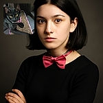 A sleek black cat with striking yellow eyes lounges comfortably on a textured cardboard scratcher, surrounded by dark wooden planks. The cat's curious gaze is fixed on the camera, its pink collar adding a pop of color to the moody scene.