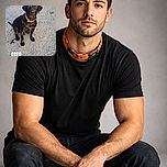 A shiny black dog with a bright orange collar sits obediently on a gravel path, looking up with soulful eyes. The photo captures the warm light of the setting sun, highlighting the dog's glossy coat. The word 'OREO' is boldly overlaid near the dog's front paws, adding a playful touch to the scene.