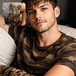 A cozy scene where a tabby cat with striking green eyes is nestled comfortably in the arms of a person wearing a white shirt, looking curiously at something off-camera. The background shows a slightly blurred kitchen area, adding a homely vibe.