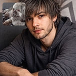 A fluffy, grumpy-looking small dog lounges on a patterned blanket on the couch, one tiny fang peeking out as if it just remembered a secret — soft fur, soulful brown eyes, and a decorative pillow and wooden furniture in the background.