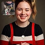 A cheerful corgi wearing a festive red harness sits proudly on a gray leather couch, flashing a big doggy smile. Behind the dog, a person in a pink sweater is taking a photo with a phone, adding a candid and cozy vibe to the living room scene. The background includes a side table with a ketchup bottle and some dishes, completing the homey atmosphere.