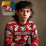 A small dog wearing a festive red and white reindeer-patterned onesie sits adorably on a gray couch with colorful pillows in the background, looking curiously at the camera.