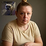 A curious French Bulldog stares directly into the camera with big, soulful eyes while sitting on a textured carpet. The cozy indoor setting includes a person's foot in a fuzzy slipper and a hint of another dog in the background, creating a warm, homey vibe.