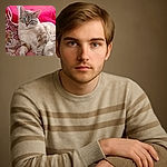 A fluffy cream-colored cat with striking blue eyes lounges elegantly on a patterned pink and red fabric, looking curiously at the camera with an air of regal relaxation.