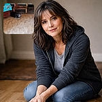 A small, grey-muzzled dog is curled on the hardwood floor near a low wooden step, peeking up at the camera with a guilty/curious expression while a bright blue blanket hangs over furniture nearby.
