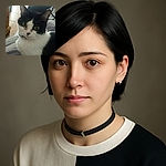 A close-up photo of a black and white cat with striking green eyes, sitting comfortably on a bed with a geometric patterned blanket in the background. The image is slightly grainy but captures the cat's curious expression well.