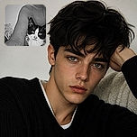 A tuxedo cat peeks out from beneath a person’s arm with the solemn expression of a tiny monochrome detective, while the tattooed arm forms an accidental cat-sized archway. The black-and-white styling makes the whole cuddle scene look dramatically cinematic.
